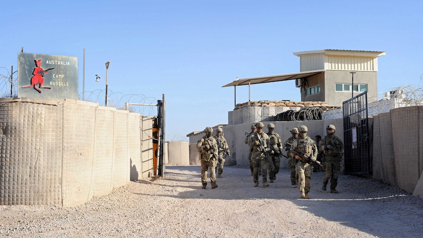 A group of soldiers, whose faces are blurred, walk down a dusty track past a sign that says "Australia, Camp Russell".