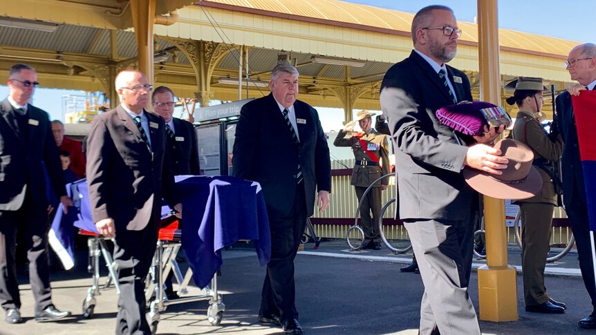 Four older men in suits wheel a coffin draped in an Australian flag along a country railway station platform.