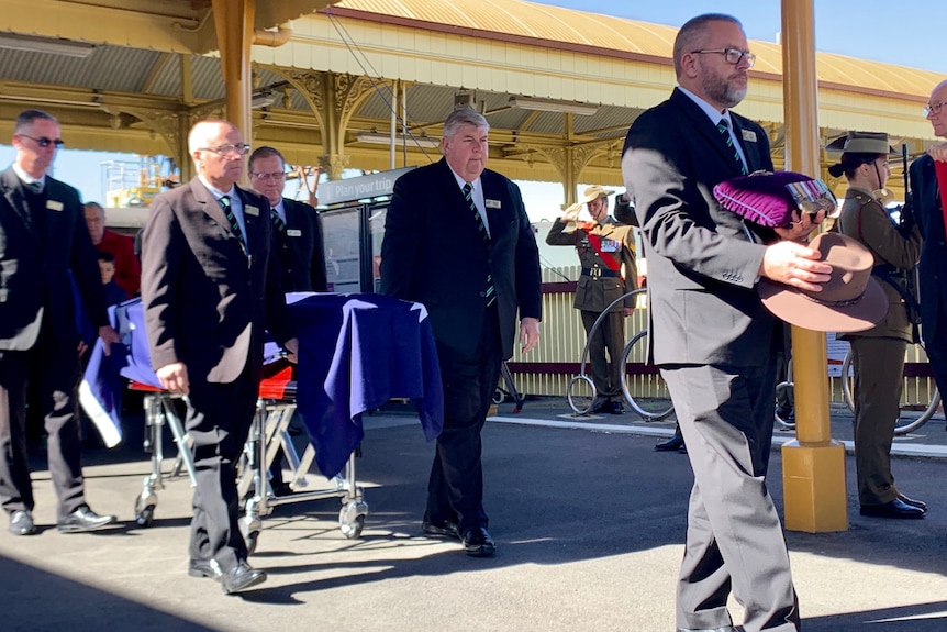 Four older men in suits wheel a coffin draped in an Australian flag along a country railway station platform.