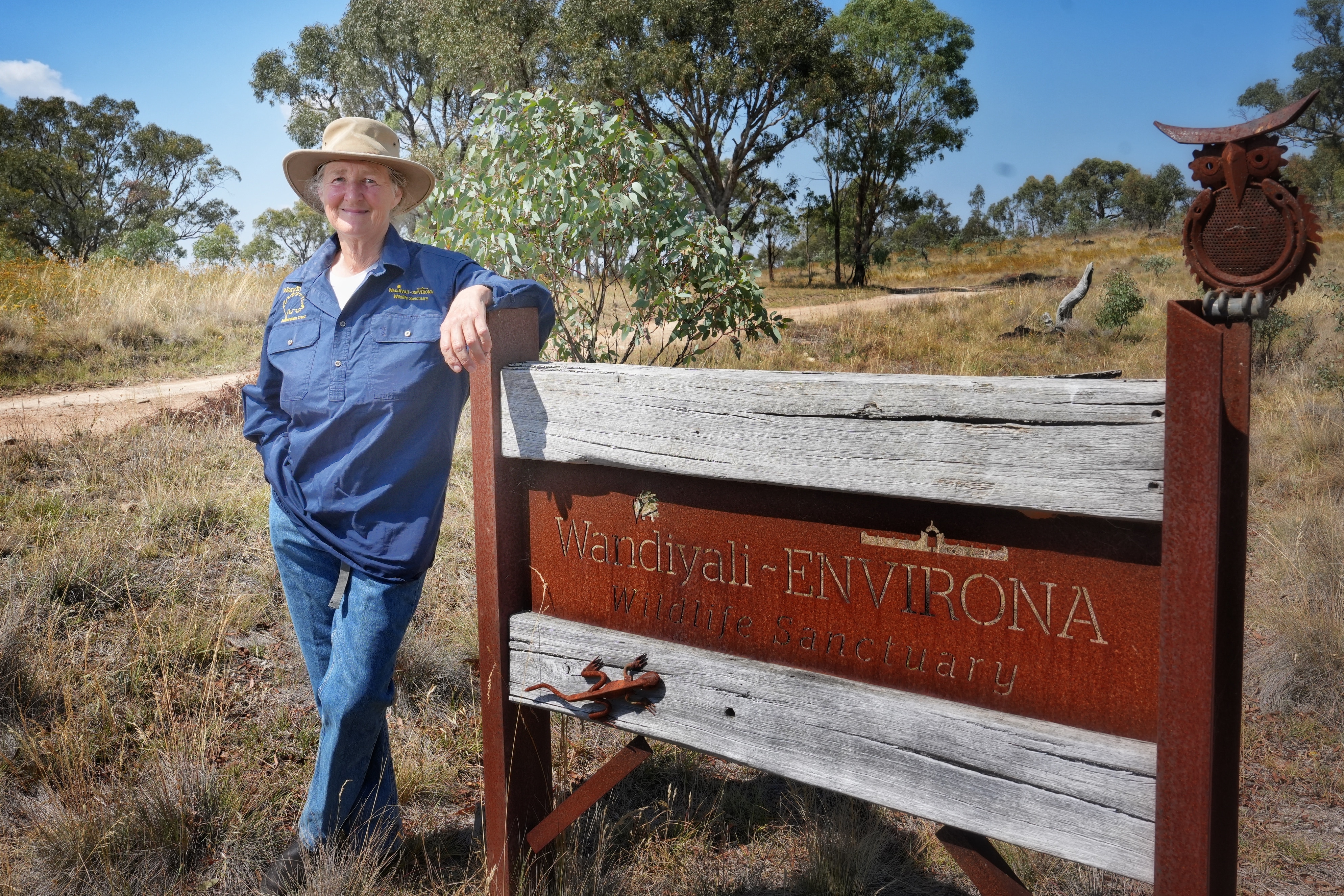 Una mujer con cabello gris se apoya contra un cartel en una propiedad de arbustos.