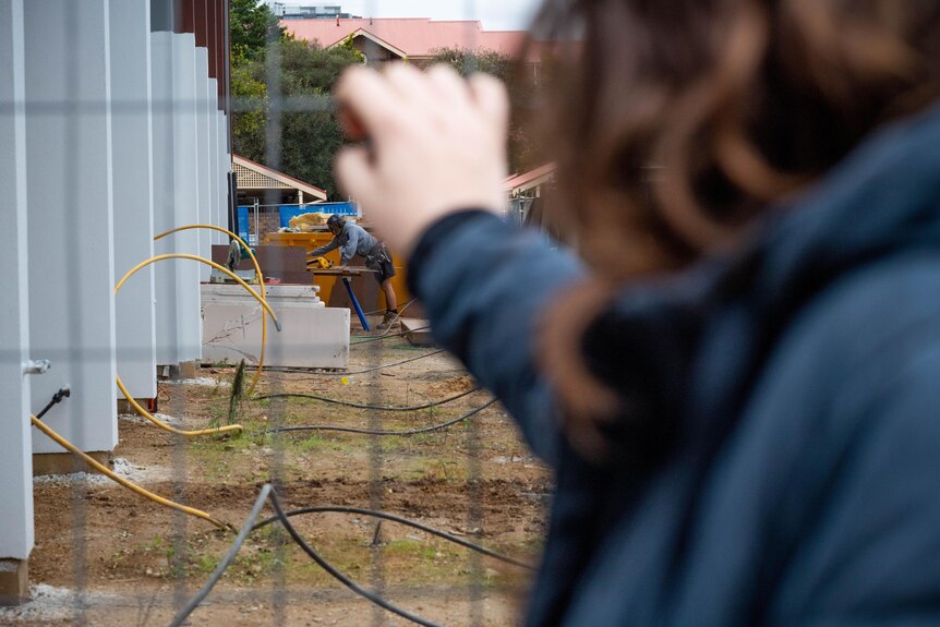 A lady standing with her arm up on a fence with a tradie in the distance 