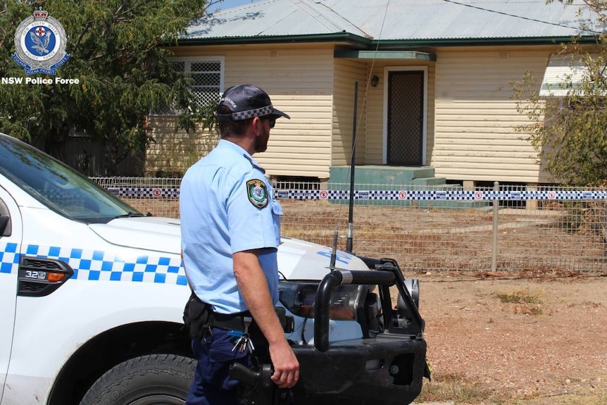 A police officer standing next to a police vehicle in front of a house with police tape around it.