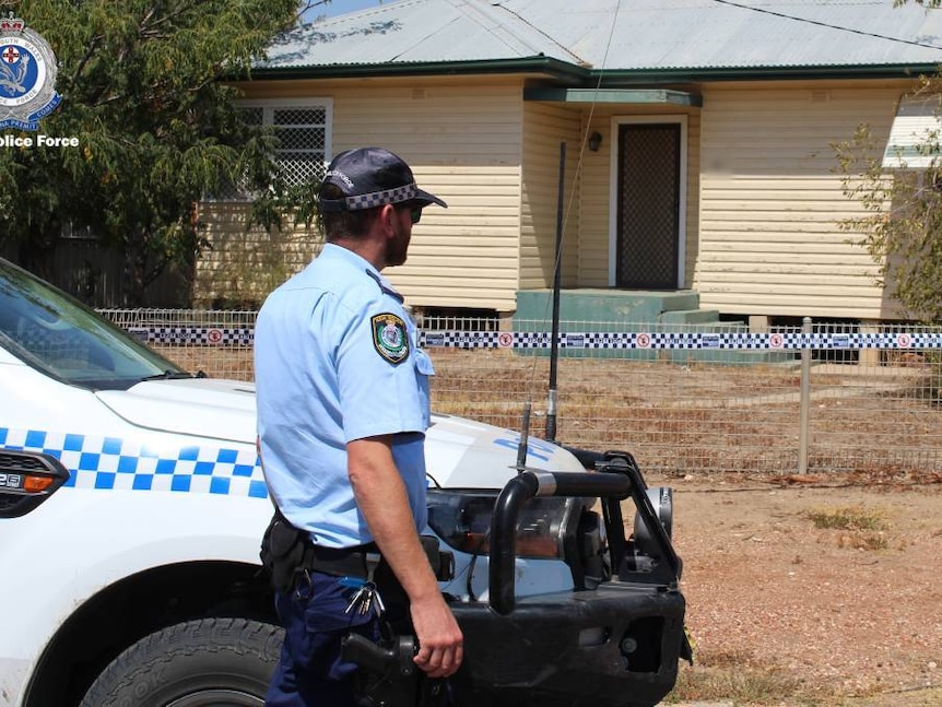 A police officer standing next to a police vehicle in front of a house with police tape around it