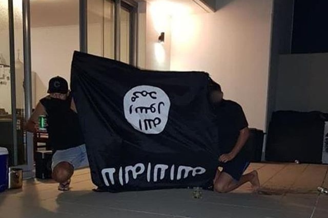 Two men with faces obscured crouch on balcony at night holding large black ISIS flag. One is holding a green beer tin