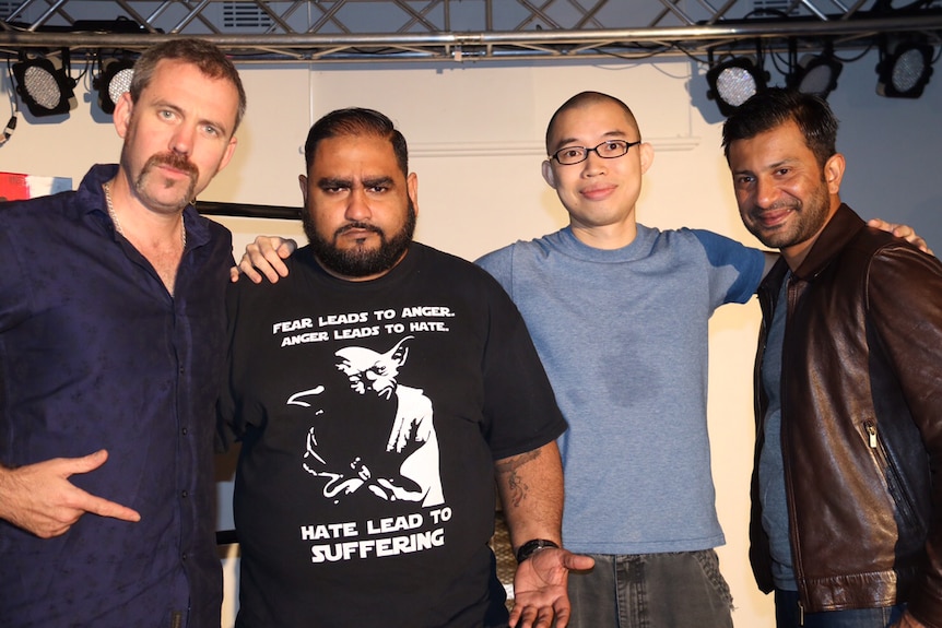 Four men pose for a photo on a stage with their arms around one another.