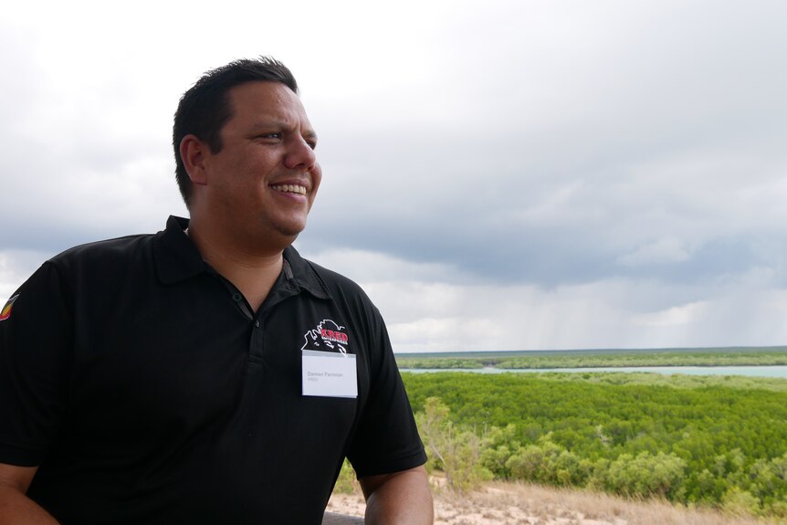 A dark skinned man wearing a black polo shirt and a name tag stands in front of a mangrove swamp on a cloudy day.