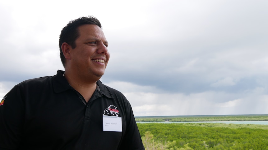 A dark skinned man wearing a black polo shirt and a name tag stands in front of a mangrove swamp on a cloudy day.