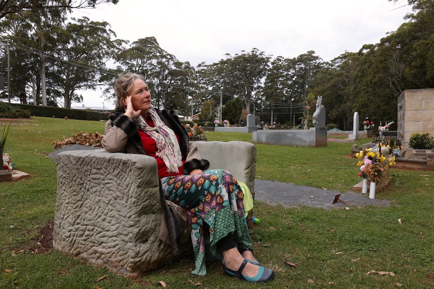 A woman sits on a tombstone in a cemetery, looking sad.