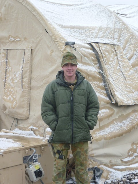 A male soldier stands outside a tent wearing winter weight army gear and cap.