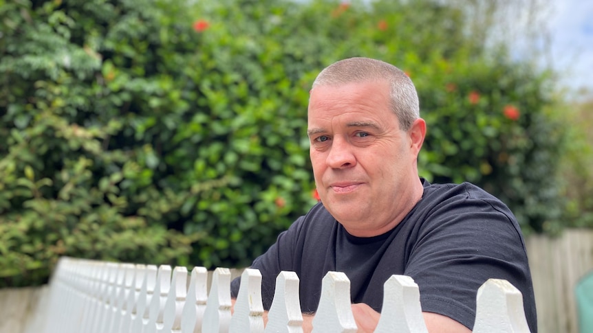 A man in a black t-shirt with close cropped grey hair stands behind a white picket fence.