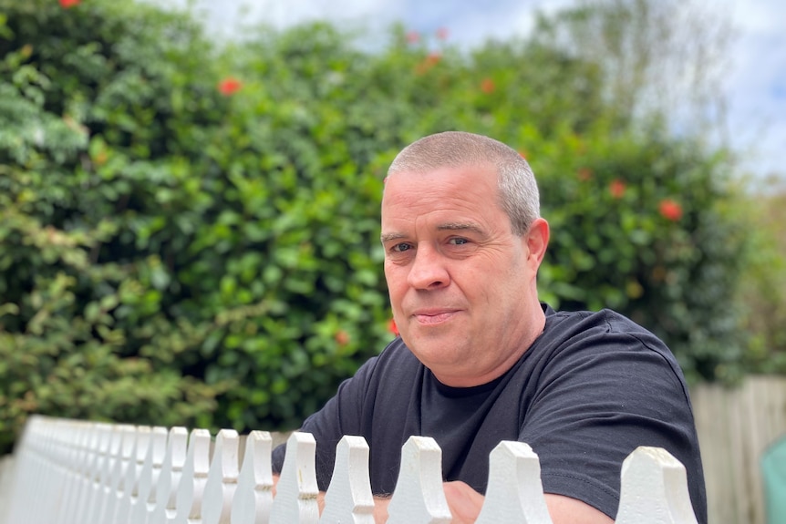 A man in a black t-shirt with close cropped grey hair stands behind a white picket fence.