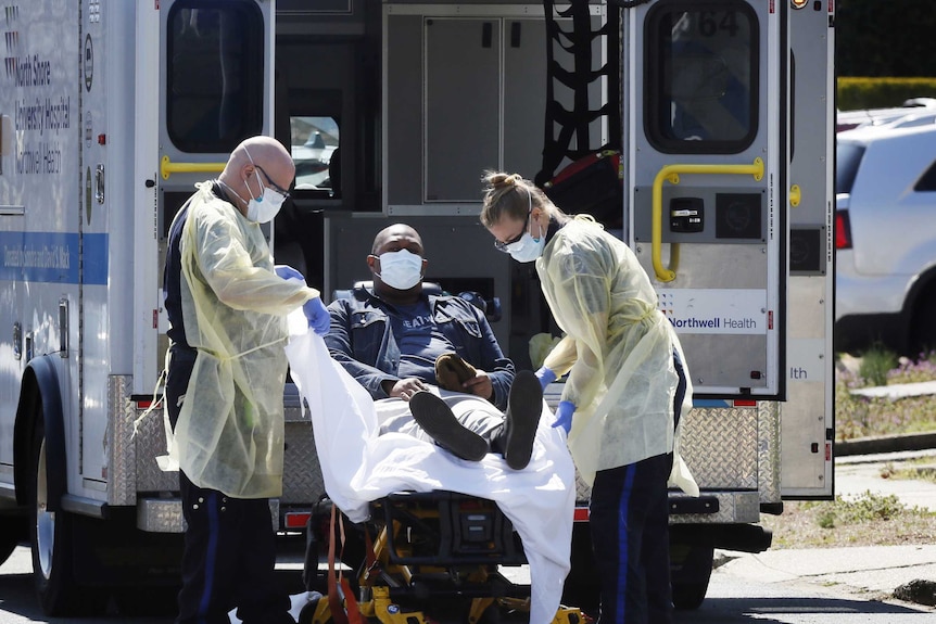 Two ambulance officers in protective gear lift a patient into the back of an ambulance.