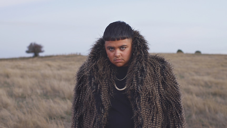 Colour image of artist Mojo Juju looking towards camera wearing feathery coat and standing in grassy plain.