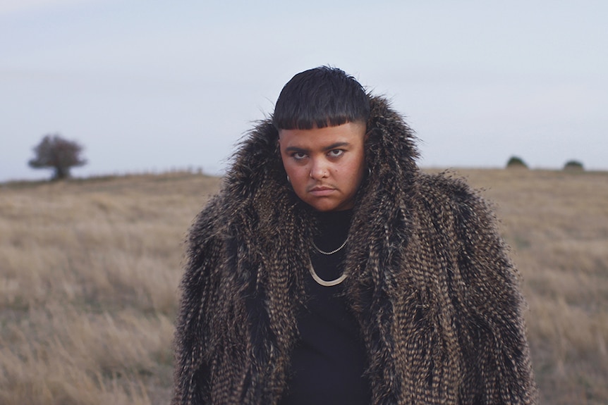 Colour image of artist Mojo Juju looking towards camera wearing feathery coat and standing in grassy plain.