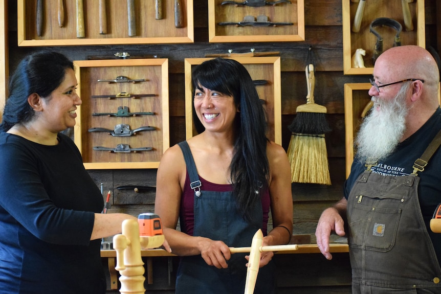 Three people talking and smiling in a workshop. Tools are on the wall. 