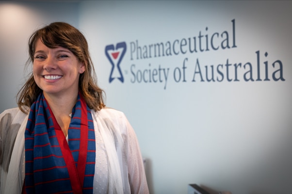 A woman, wearing a white lab coat, smiles in front of a pharmacy society sign