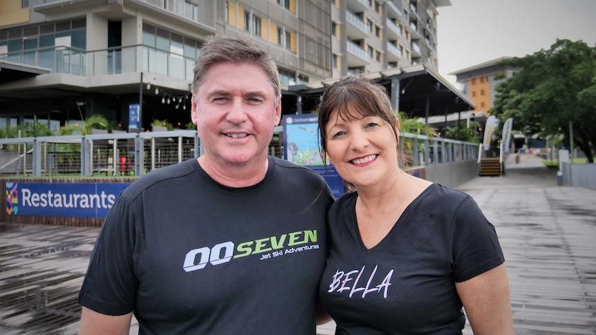 Man wearing black 00Seven T-shirt and with woman wearing black Bella Tshirt standing with high-rise apartments behind.