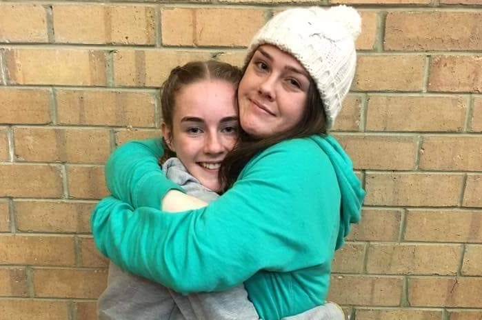 Two girls in turquoise and grey jumpers hug as they pose for a photo.