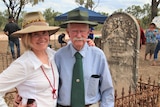 Woman in white shirt with elderly man in blue shirt standing in front of a gravestone 