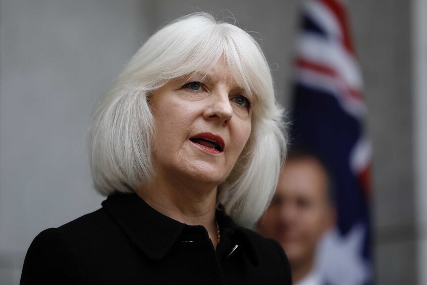 Mid shot of Morgan, wearing a black top, grey hair, standing in front of out-of-focus Australian flag.