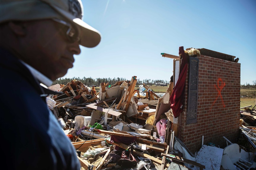 A man stands next to a pile of rubble, and the partial brick wall of a house.