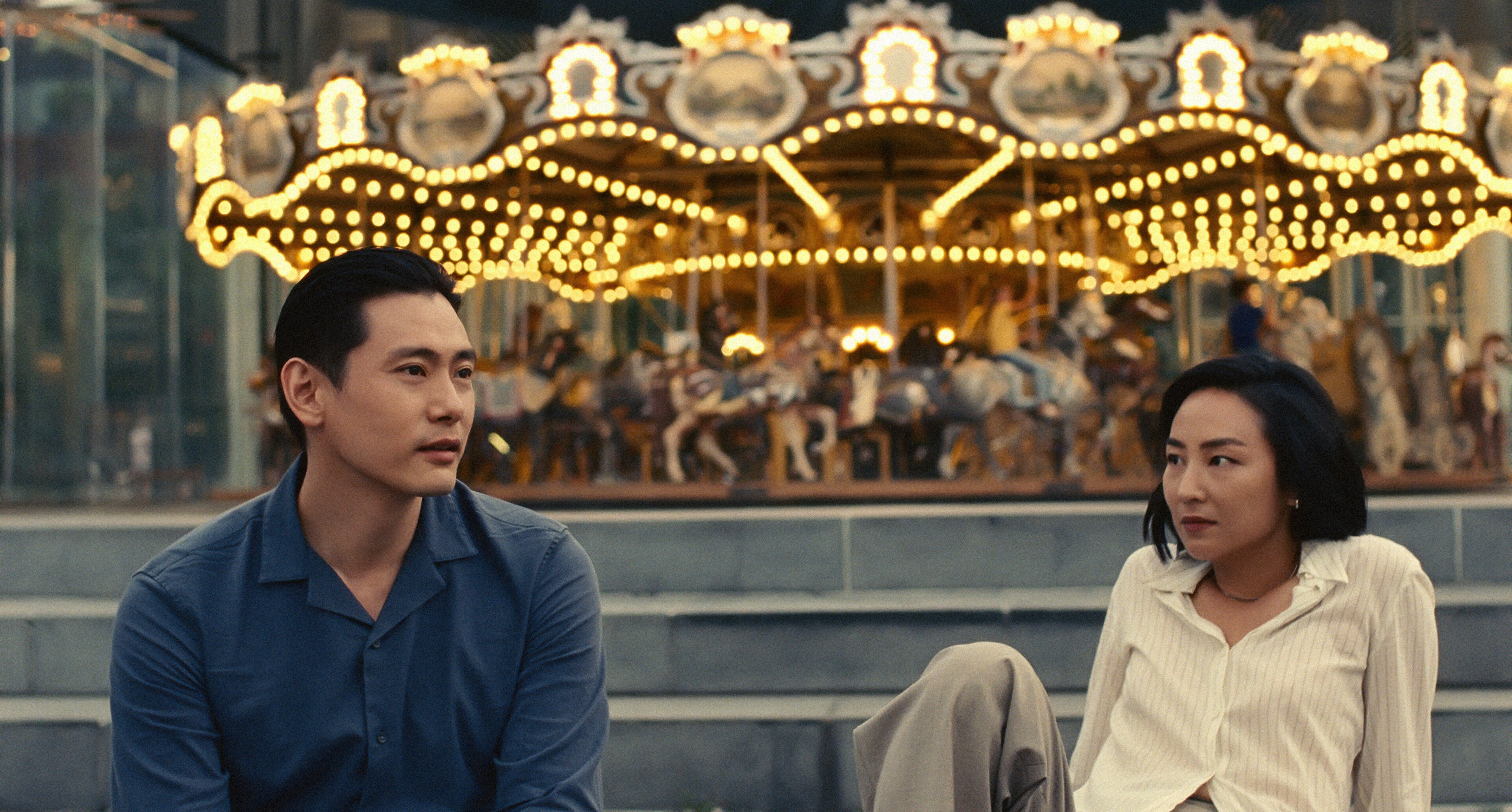 A Korean man with dark hair wears a navy shirt and laughs, walking beside a Korean woman with short dark hair and a white blouse