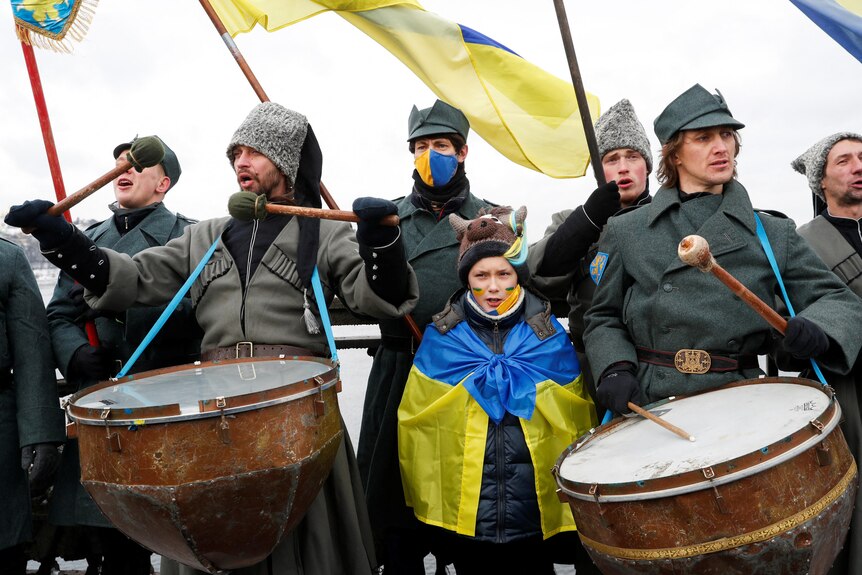 A little girl wrapped in a Ukrainian flag stands among a group of men playing drums