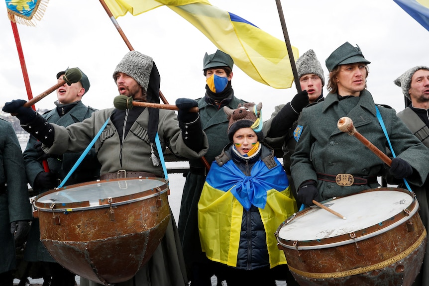 A little girl wrapped in a Ukrainian flag stands among a group of men playing drums