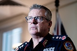 A serious older man, spiky, short grey hair, glasses, uniform with police written on it, Australian flag behind, mike in front.