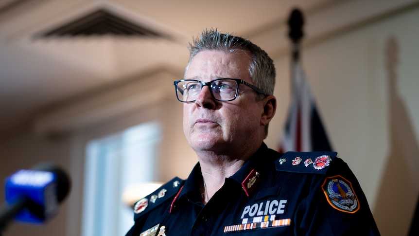 A serious older man, spiky, short grey hair, glasses, uniform with police written on it, Australian flag behind, mike in front.