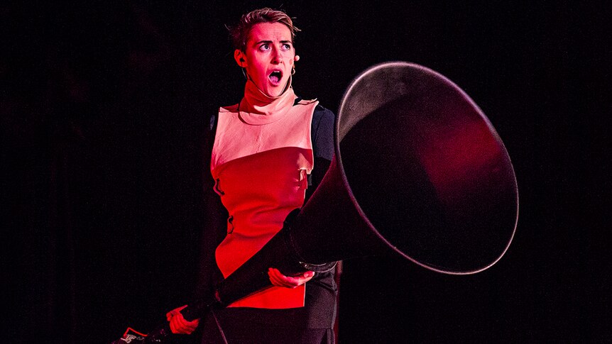 The singer stands on stage, lit in red, mouth agape, holding an enormous megaphone-like object.
