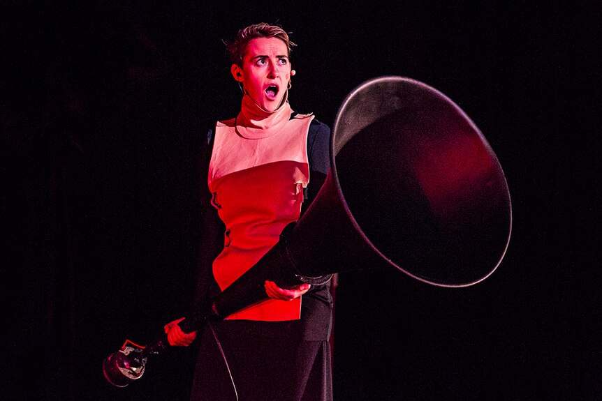 The singer stands on stage, lit in red, mouth agape, holding an enormous megaphone-like object.