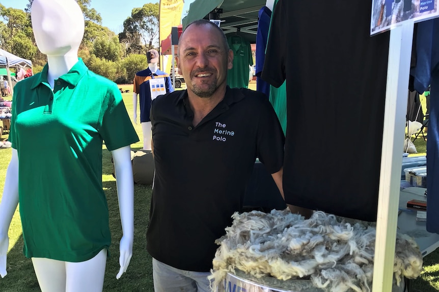 Man standing wearing a black polo shirt standing outside at a market stall, next to a mannequin with green polo shirt.