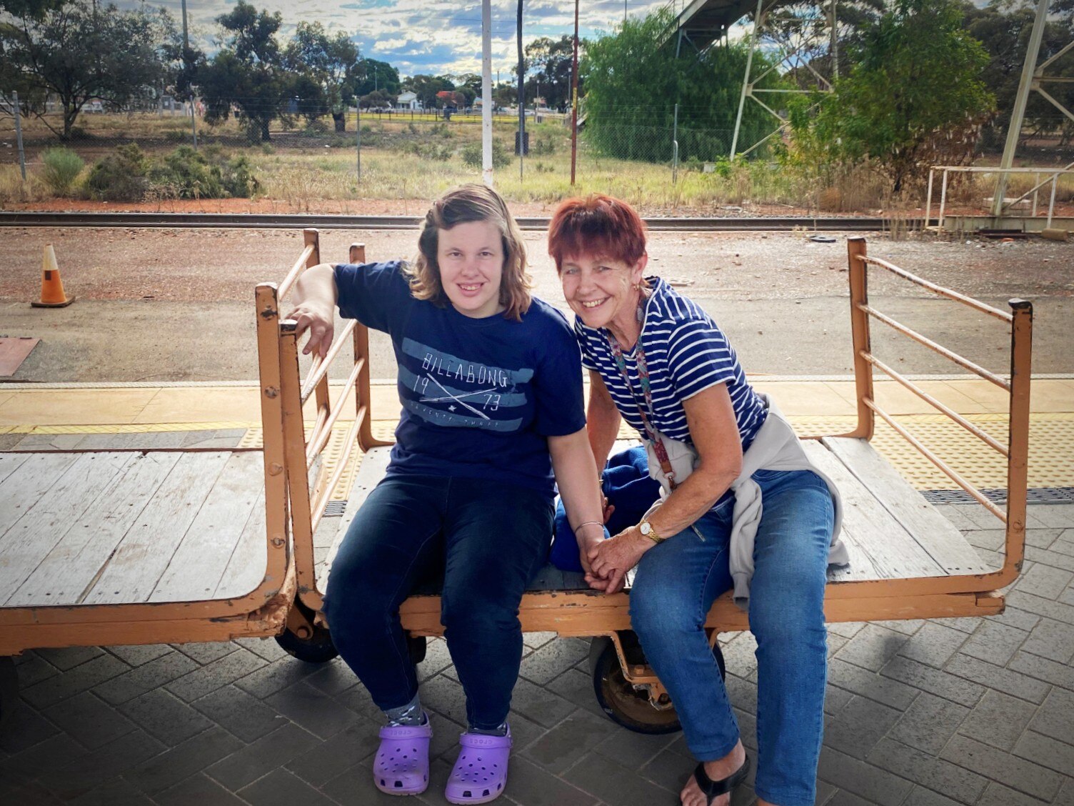 A younger woman with shoulder length mousy blonde hair and an older woman, short red hair, sitting together on a cart, smiling.