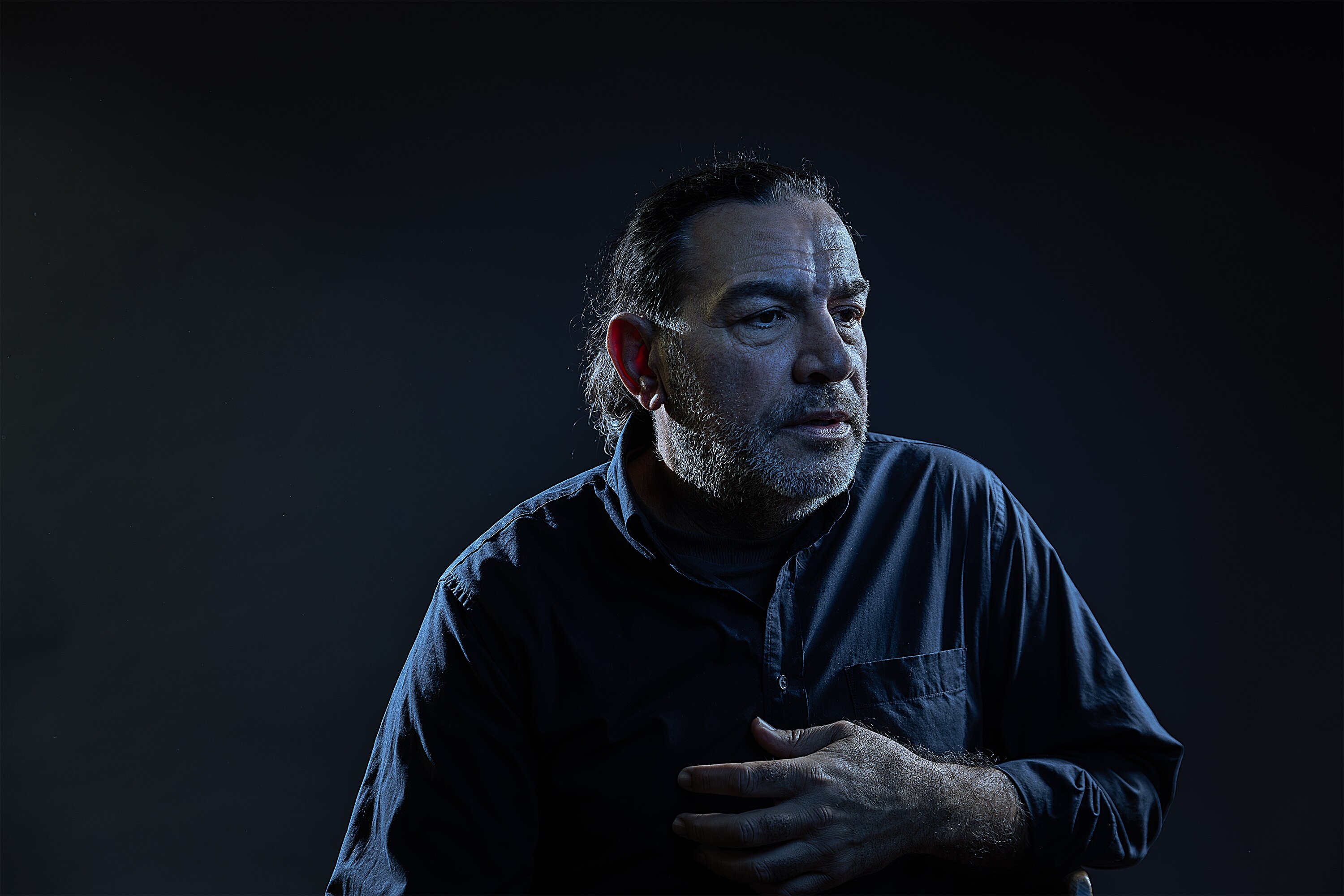 Against a dark background, Khaled Sabsabi looks off to side, wearing a dark shirt with shoulders back hand on chest.