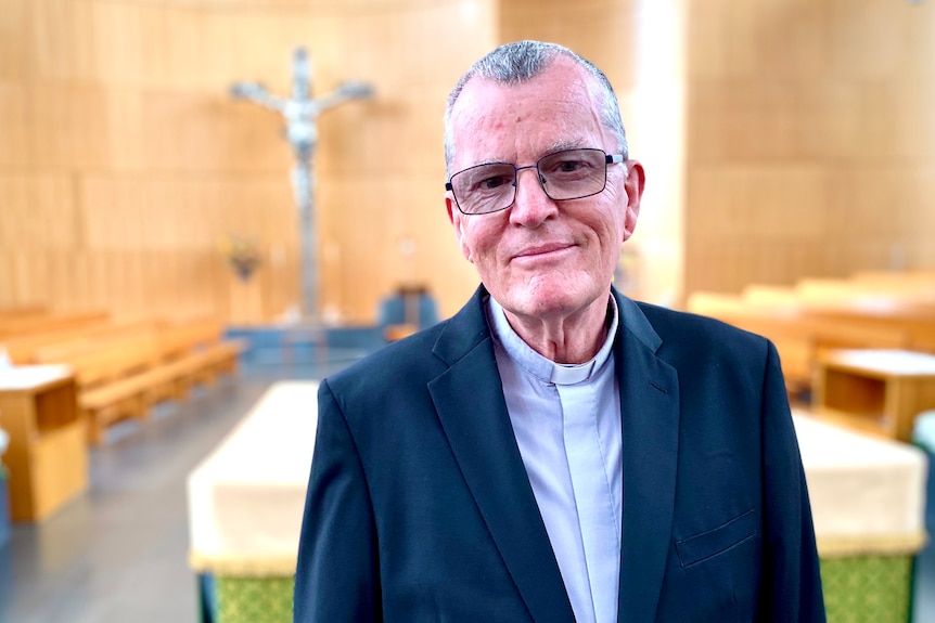 A man wearing a priest's collar smiles at the camera with a crucifix behind. 