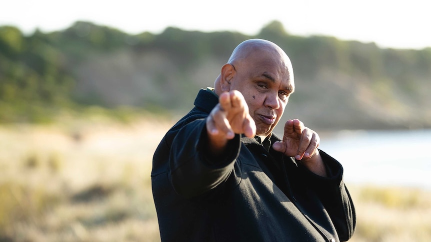 Kutcha Edwards, wearing a black shirt, is turned to the side, face looking forward, pointing with two fingers on each hand