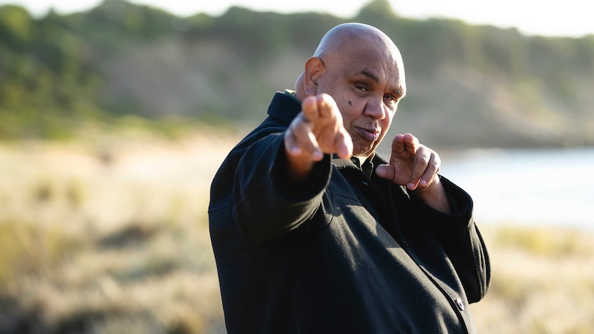 Kutcha Edwards, wearing a black shirt, is turned to the side, face looking forward, pointing with two fingers on each hand