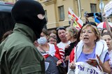 A woman standing in the front of a large group of protesters argues with a police officer wearing a balaclava.