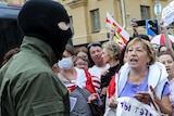 A woman standing in the front of a large group of protesters argues with a police officer wearing a balaclava.