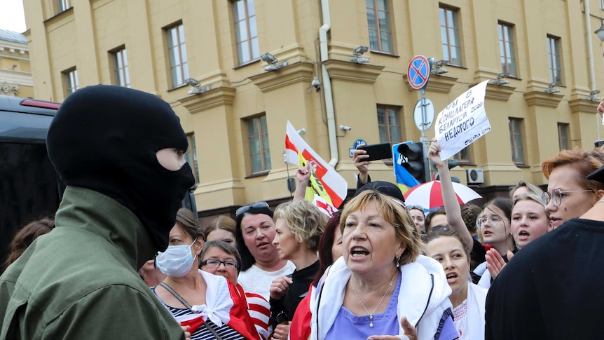 A woman standing in the front of a large group of protesters argues with a police officer wearing a balaclava.