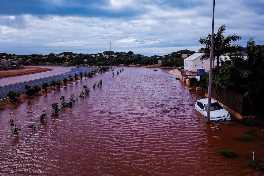 Red-brown flood waters cover a street, with a white car to the right. 