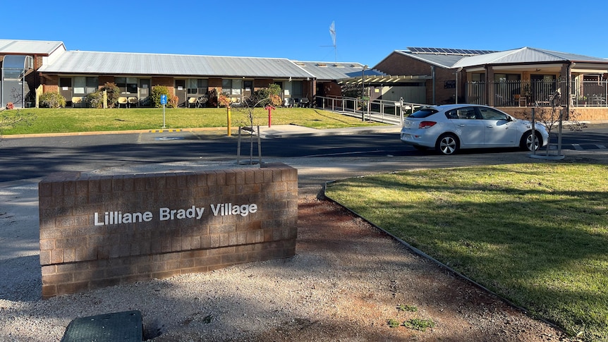 A low building, spread across the lawn, grey, steel roof, sign saying Liliane Brady Village, a white car parked outside.