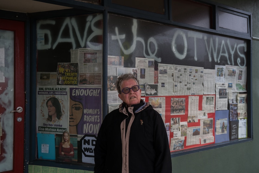 A woman stands in front of a shop front with newspaper windows which say 'Save the Otways' above the window.