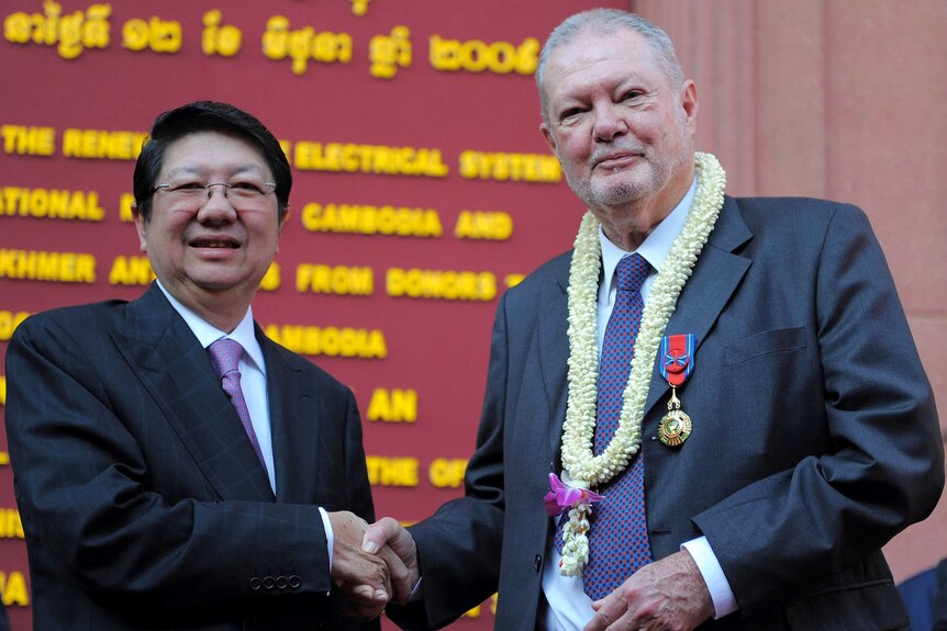 Two men in suits, one wearing a lotus flower wreath around his neck, post for a photo shaking hands.