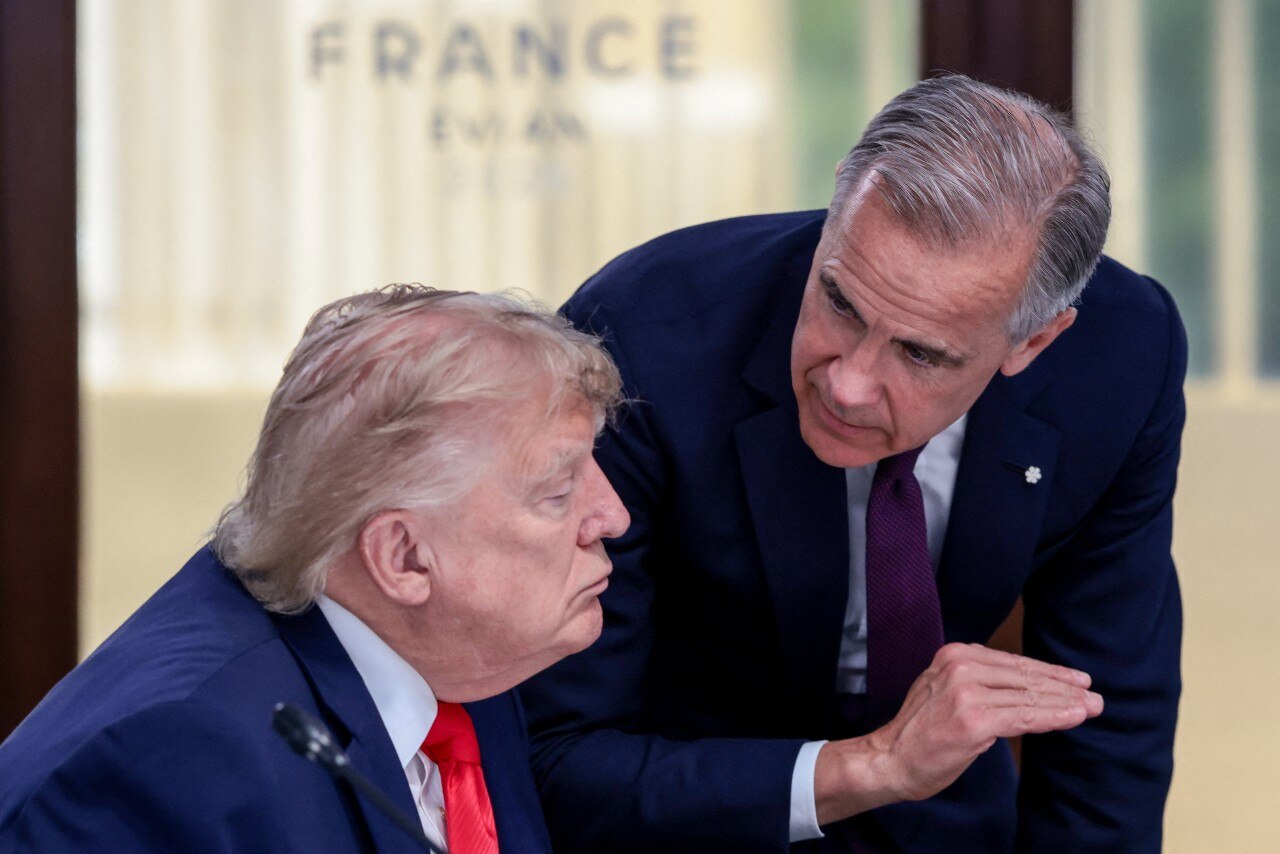 A man in a suit standing and talking to a man in a suit who is sitting.