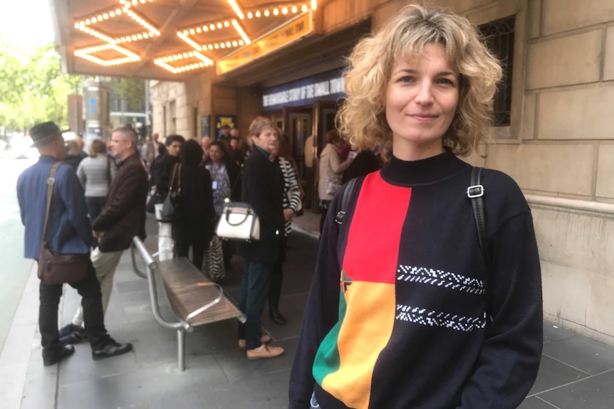 Xani Kolac stands on the street, in front of a theatre where people are lining up to get inside.