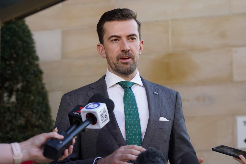 A picture of Zak Kirkup wearing a suit and green tie talking to reporters.