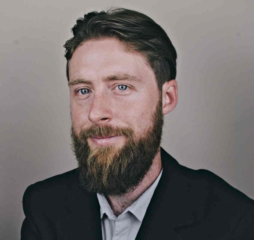 Uma foto aproximada de um homem barbudo vestindo jaqueta e camisa.