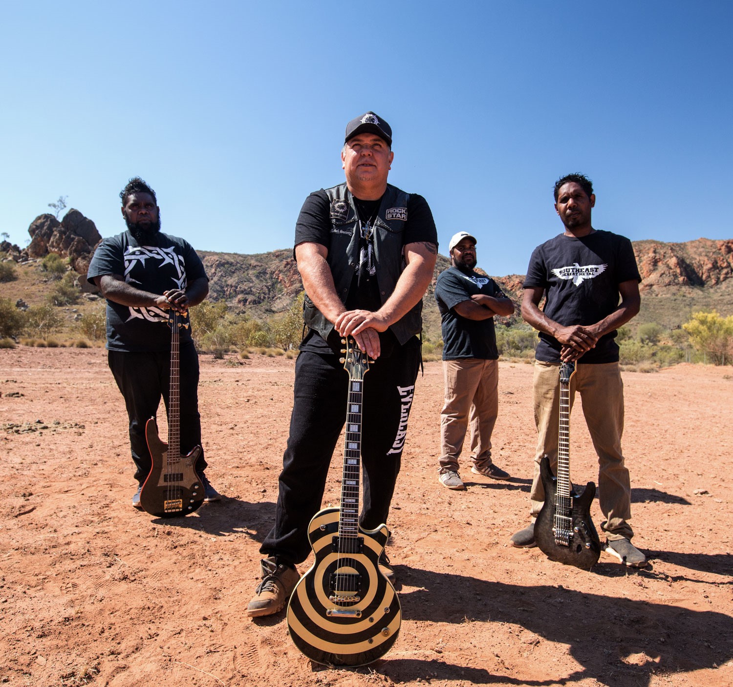 four men standing with their guitars outside surrounded by sand and rock mountains